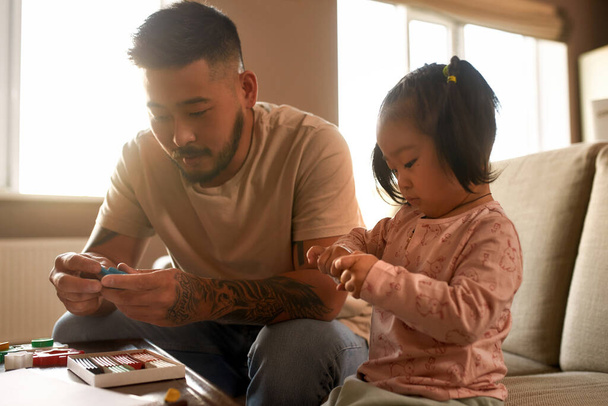 Focused father and daughter sculpting with plasticine at table at home. Asian man and little girl spend time together. Domestic entertainment and leisure. Family relationship. Parenting and fatherhood - 写真・画像