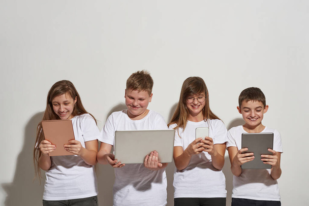 Smiling european boys and girls using variety digital gadgets. Friends with smartphone, digital tablets and laptop. Modern youngster lifestyle. Gadget addiction. White background in studio. Copy space - Photo, Image