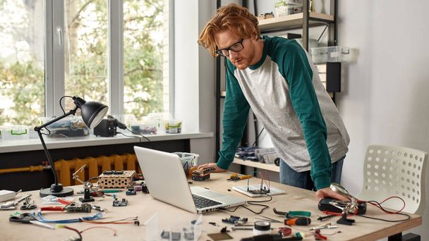 Male IT technician or engineer watching laptop connecting with microchip in office. Young focused caucasian man wearing glasses at table with tools and components. Modern technology and innovation - Fotoğraf, Görsel