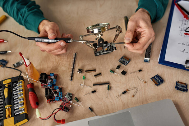 Top view of partial male technician or engineer soldering microchip with soldering iron under magnifying glass at table with laptop, variety technical tools and components. Technology and innovation - Foto, immagini