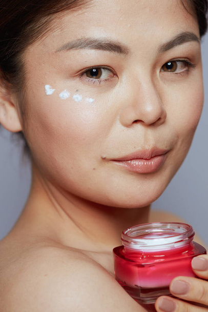 young woman with facial cream jar and eye cream on face against blue background. - Foto, afbeelding