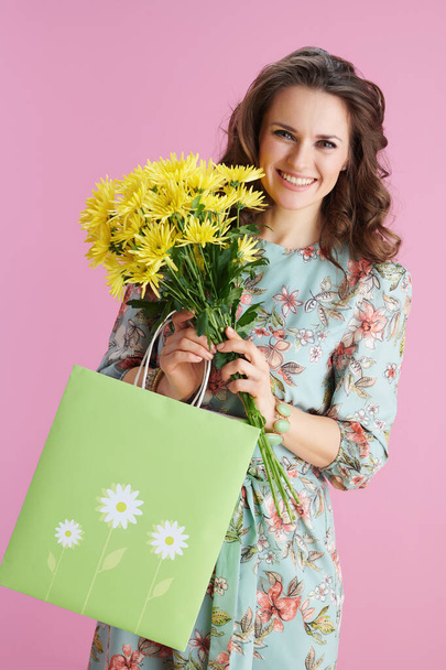 smiling elegant 40 years old woman with long wavy brunette hair with yellow chrysanthemums flowers and green shopping bag against pink background. - Foto, afbeelding