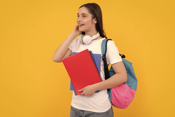 Sırt çantası ve kulaklıklı mutlu genç kız sarı arka plan ve bilgi üzerine çalışma kitabı tutuyor.. - Fotoğraf, Görsel
