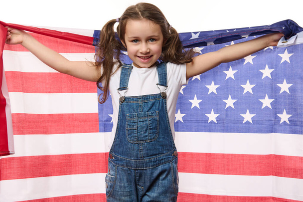 Niño caucásico feliz, linda niña vistiendo un mono de mezclilla azul, sonriendo una hermosa sonrisa dentada, mirando a la cámara, sosteniendo la bandera estadounidense en sus manos extendidas. 4 de julio - Día de la Independencia - Foto, Imagen