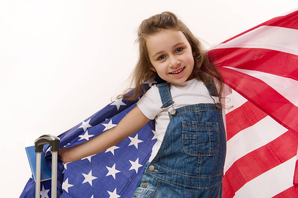 Happy baby girl wearing blue denim overalls with flag of USA, passport, boarding pass and suitcase, isolated over white background with free space for promotional text. Concept of travel, immigration - Photo, Image