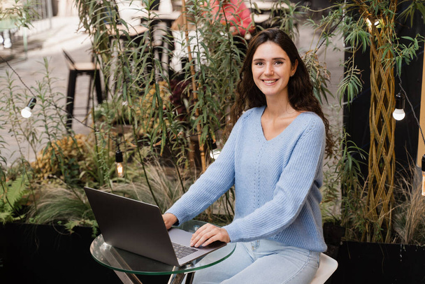 Atraktivní dívka s laptopem sedí v kavárně venku a chatuje s přáteli a kolegy. Mladá žena pracuje na notebooku vzdáleně venku v restauraci - Fotografie, Obrázek