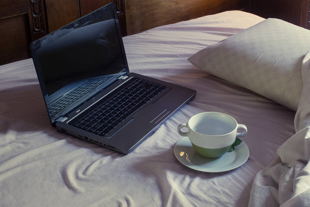Uma caneca vazia em um pires sujo ao lado de um laptop aberto em uma cama de quarto não feita com cobertores enrugados. Quarto bagunçado. - Foto, Imagem
