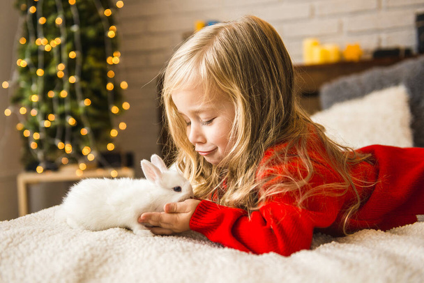 A little girl with a cute white rabbit is lying on the bed. Rabbit symbol of 2023 - Zdjęcie, obraz