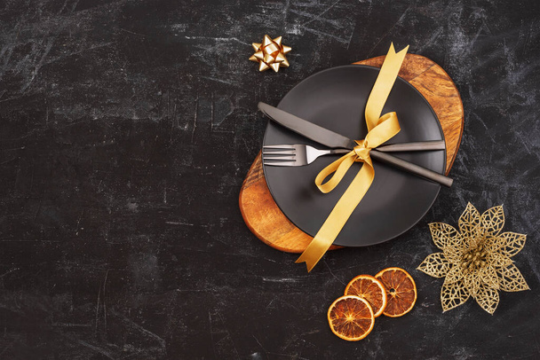 Table noire avec une assiette sur planche à découper, fourchette et couteau avec arc doré, décorations de Noël et tranches d'orange. Vue du dessus, plan, espace de copie. - Photo, image