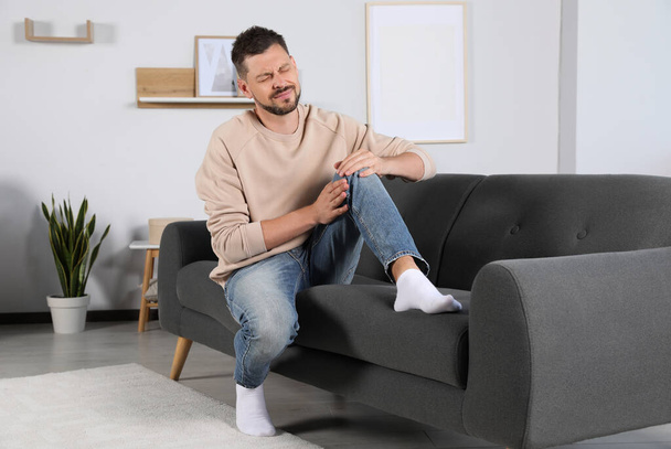 Man suffering from knee pain on sofa at home - Fotoğraf, Görsel