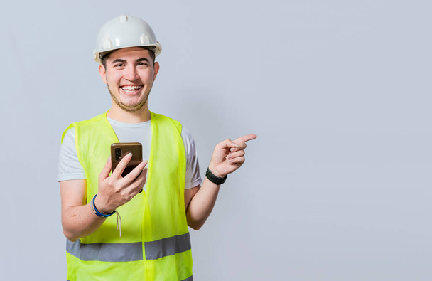 Happy Civil engineer holding cellphone and pointing a promo. Happy engineer holding phone and pointing to the side isolated. Young construction engineer holding phone and pointing at promo - Foto, Imagem