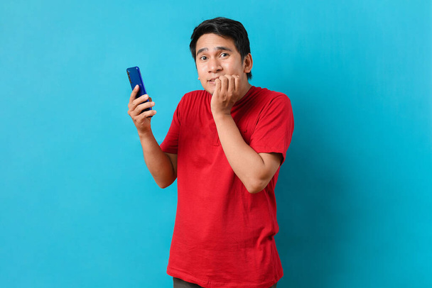 Young Asian man holding a mobile phone with shocked expression, isolated on blue background - Zdjęcie, obraz