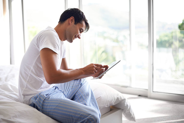 Early morning browsing. a handsome young man using a tablet on the edge of his bed - Foto, afbeelding