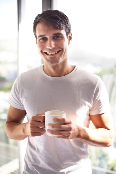 Er gaat niets boven een verse kop koffie in de ochtend. Portret van een knappe jongeman die koffie drinkt terwijl hij bij het raam van zijn slaapkamer staat - Foto, afbeelding