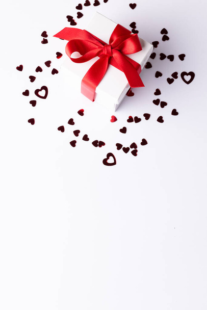 Vertical of white gift box with red ribbon on heart confetti, on white background with copy space. Valentine's day, love, romance and celebration concept. - Foto, afbeelding