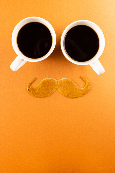 Composition of fake moustache and cups of coffee on orange background with copy space. Fake moustache day, party, celebration and costume concept. - Foto, Bild