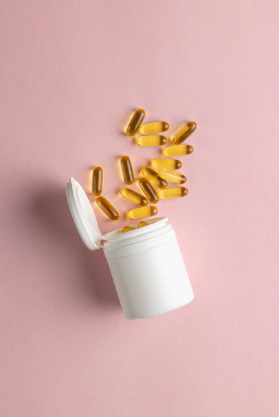 Vertical composition of white pill box spilling oil capsules on pink background with copy space. Medicine, medical services, healthcare and health awareness concept. - Fotografie, Obrázek