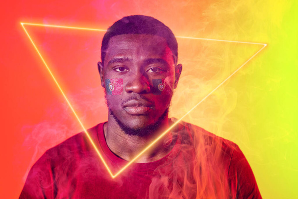 Portrait of african american male soccer player with portuguese flag on face by illuminated triangle. Copy space, confident, composite, sport, soccer, competition, neon, illustration, smoke. - Фото, изображение