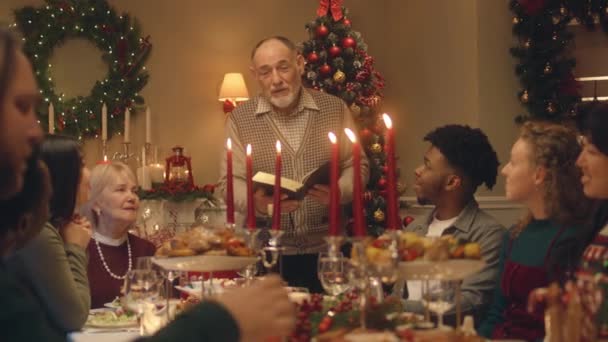 Drandfather lee la Biblia en la cena familiar de Navidad. Feliz gran familia rezando antes de celebrar la Navidad o Año Nuevo 2023. Se sirve mesa de vacaciones con deliciosa comida y velas. Vacaciones de invierno. - Imágenes, Vídeo
