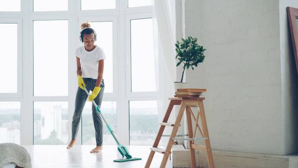 African American girl is doing housework in nice tidy flat, young woman is mopping floor and having fun listening to music, dancing and singing. Home, people and cleanliness concept. - 写真・画像