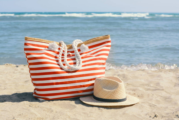 Stylish striped bag with straw hat on sandy beach near sea - Zdjęcie, obraz