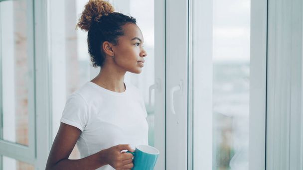 Smiling young African American woman is drinking tea standing near the window and looking outside enjoying leisure time at home. Apartments, drinks and people concept. - Foto, imagen