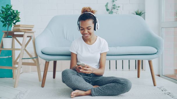 Chica bonita está jugando juego en la pantalla táctil del teléfono inteligente y sonriendo mientras escucha música con auriculares sentados en el suelo en casa. Tecnología, millennials y concepto divertido
. - Foto, imagen