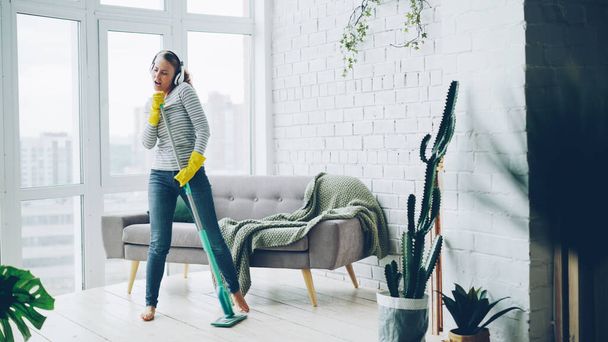 Happy young housewife is dancing and singing during housecleaning, she is listening to music in headphones and mopping floor having fun. Housework and technology concept. - Photo, image