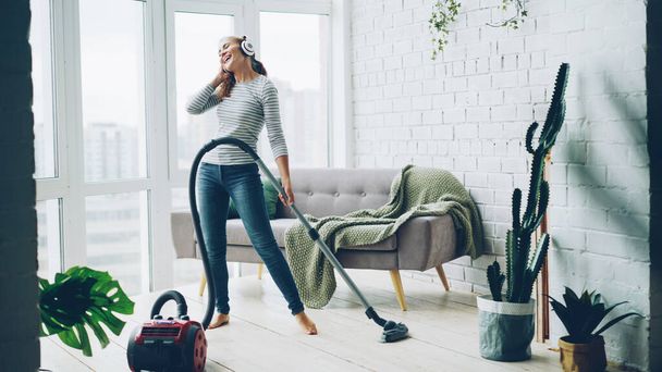 Beautiful young woman is hoovering the floor at home using modern vacuum cleaner and listening to music with headphones, dancing and singing. Housework and technology concept. - Fotó, kép