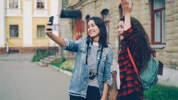 Cheerful girls foreign travelers are taking selfie using smartphone standing outdoors and posing with hand gestures showing v-sign and heart with fingers and laughing together. - Photo, image