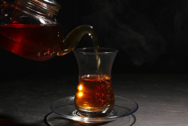 Pouring traditional Turkish tea from pot into glass on black table - Foto, Imagen