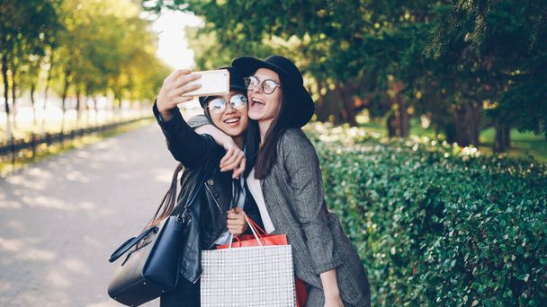 Excited female friends are taking selfie using smartphone and holding shopping bags with purchases on sunny autumn day. Modern technology and youth lifestyle concept. - Foto, afbeelding