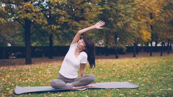 Beautiful girl is concentrated on yoga practice sitting on mat in park in lotus position and bending sideways raising arm. Healthy lifestyle, sports and recreation concept. - Foto, afbeelding