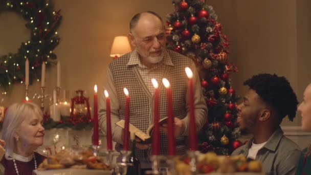 Drandfather leest de bijbel over familie kerstdiner. Grote divers gezin bidden voor Kerstmis of Nieuwjaar 2023 te vieren. Geserveerd vakantietafel met heerlijke maaltijd en kaarsen. Wintervakantie. - Video