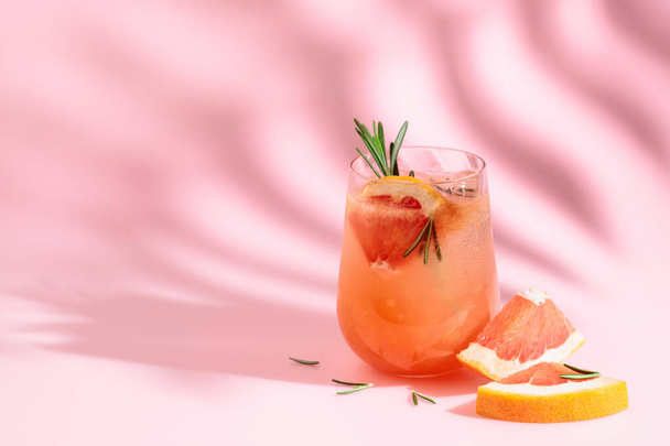 Summer cocktail with grapefruit, rosemary, and ice. Drink on pink background with palm leaf shadow. Summer, tropical, fresh cocktail concept. - Zdjęcie, obraz