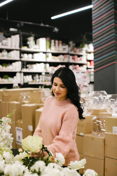 A young brunette woman in a pink sweater buys artificial flowers for the interior in the store. - Foto, afbeelding