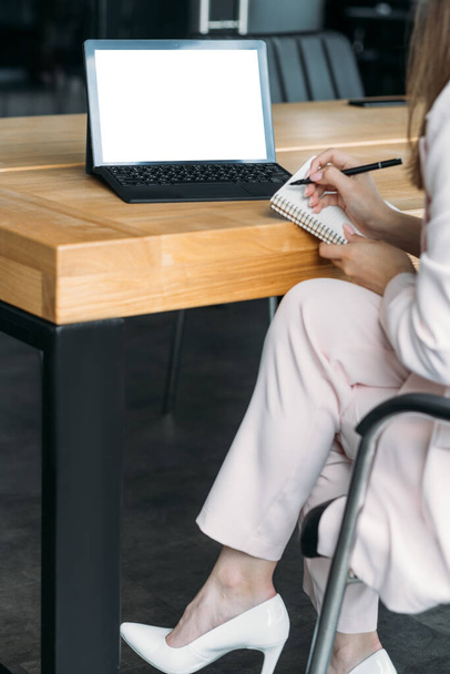 Online meeting. Computer mockup. Digital technology. Unrecognizable elegant woman sitting desk with laptop blank screen making notes notebook in light room interior. - Photo, image