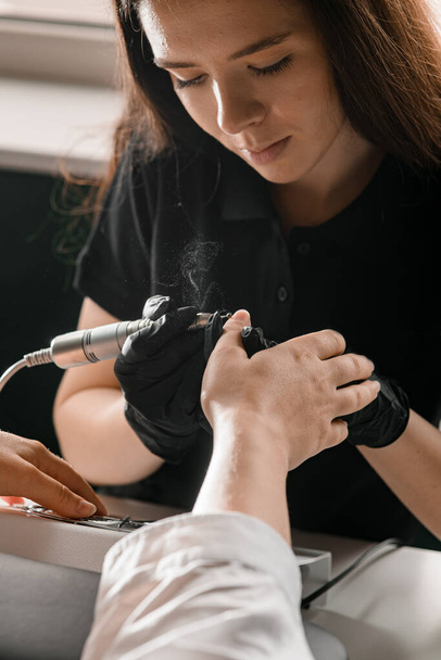 Woman manicurist in black gloves holds professional cuticle remover device and fails female hands of client. Personal care and cosmetics. Close-up. Hardware manicure. - Photo, image