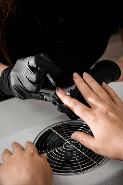 Manicurist in black gloves is applying transparent nail base gel on female finger nails. Nail care concept. - Foto, Imagen