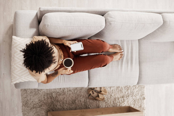 Draufsicht, entspannen und schwarze Frau mit Telefon auf dem Sofa Kaffee trinken und in den sozialen Medien, Internet und online. Kommunikation, Technik und Mädchen mit Mockup-Bildschirm auf Smartphone für mobile App zu Hause. - Foto, Bild