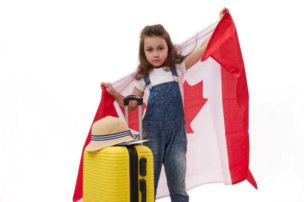 Linda niña en camiseta blanca y overoles de mezclilla azul, llevando bandera de Canadá, sonríe mirando a la cámara mientras posa con una maleta amarilla sobre fondo blanco con espacio libre para texto publicitario - Foto, Imagen