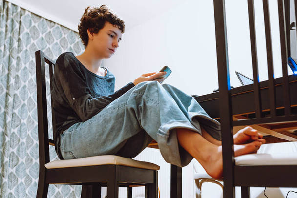 Teenager-Alltag zu Hause. Teenager-Mädchen mit braunen kurzen Haaren sitzen mit überkreuzten Beinen im heimischen Esszimmer, nutzen Smartphone, chatten oder lesen SMS. Blick von unten nach oben - Foto, Bild