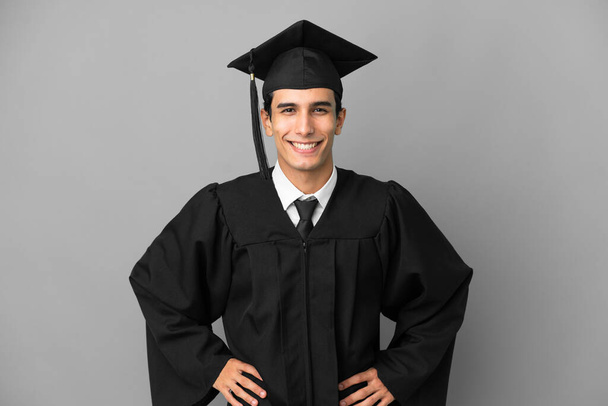 若いアルゼンチンの大学の卒業生は、腰と笑顔で腕をポーズ灰色の背景に孤立 - 写真・画像