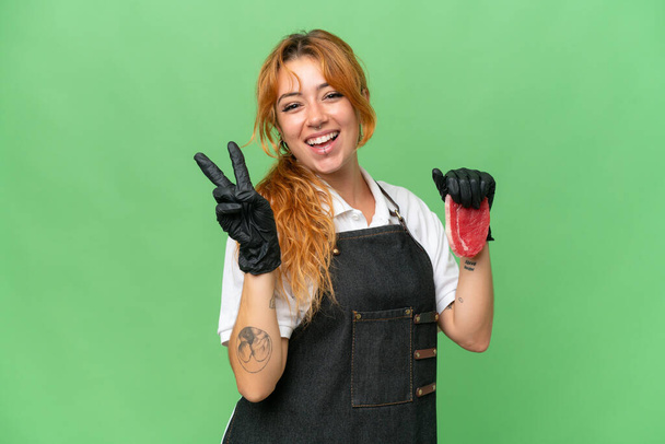Carnicero mujer caucásica usando un delantal y sirviendo carne fresca cortada aislado en pantalla verde croma clave fondo sonriendo y mostrando signo de victoria - Foto, Imagen
