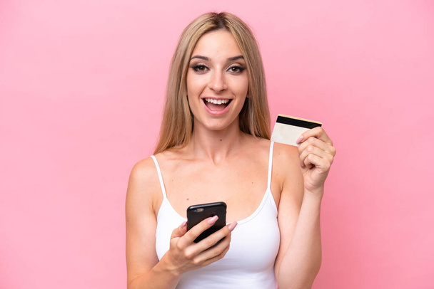 Красива блондинка ізольована на рожевому фоні, купуючи з мобільним телефоном і тримаючи кредитну картку з здивованим виразом
 - Фото, зображення