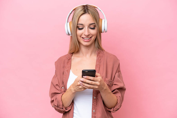 Pretty blonde woman isolated on pink background listening music and looking to mobile - 写真・画像