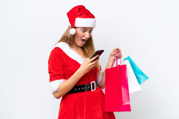 Young caucasian woman dressed as mama noel isolated on white background holding shopping bags and writing a message with her cell phone to a friend - Φωτογραφία, εικόνα