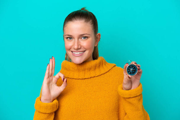 Молодая белая женщина держит компас изолирован на синем фоне показывая знак ОК с пальцами - Фото, изображение