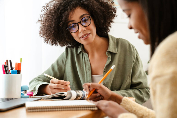 Black girl writing in exercise book while doing homework with her mother at home - Foto, Bild