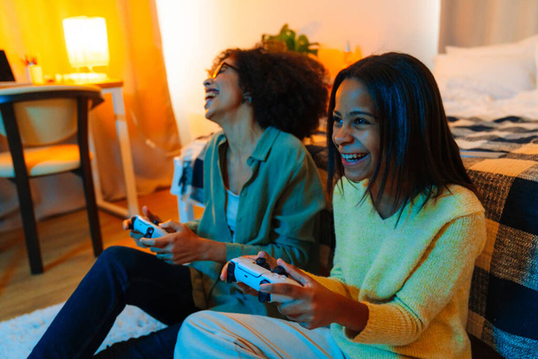 Happy african american woman and her teenage daughter playing video game with controllers at home - Φωτογραφία, εικόνα